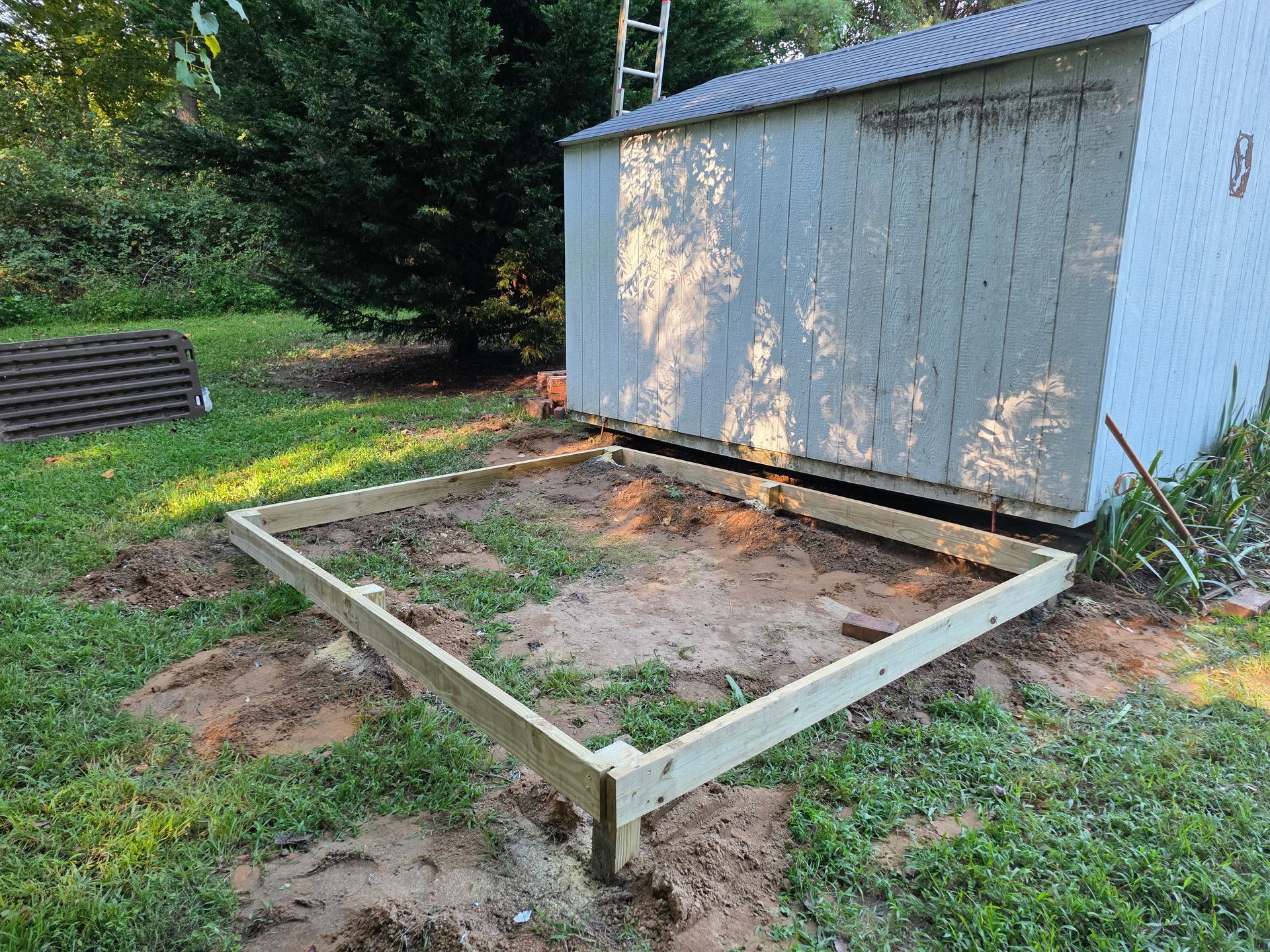 Wooden frame foundation for a shed on grass and dirt. Light blue shed in background.