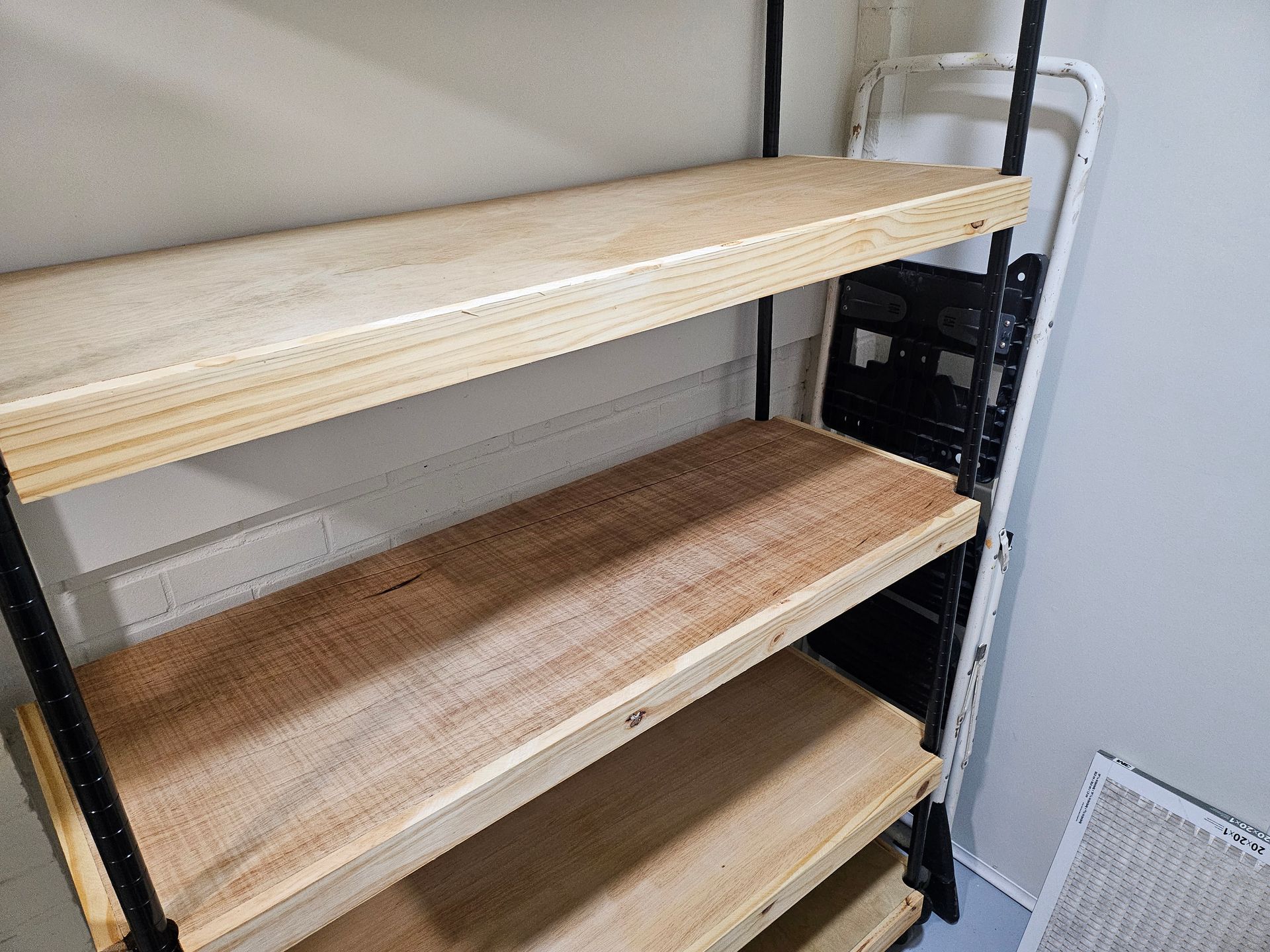 Black metal shelving unit with three wooden shelves against a white wall; a folding ladder is visible.