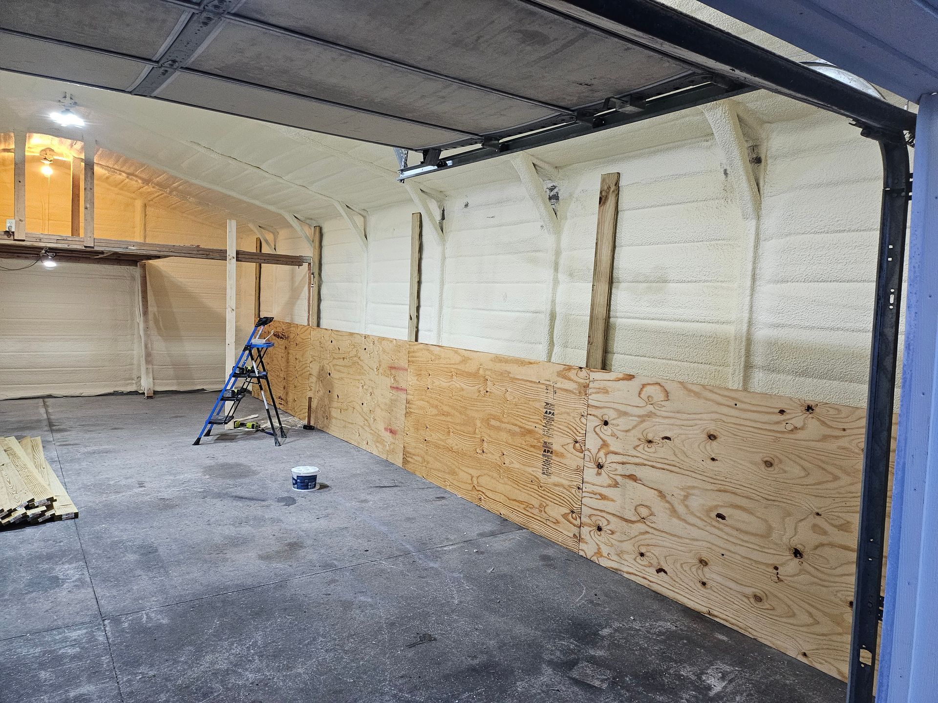 A person on a ladder works on a plywood wall inside a garage; the walls are insulated with white foam.