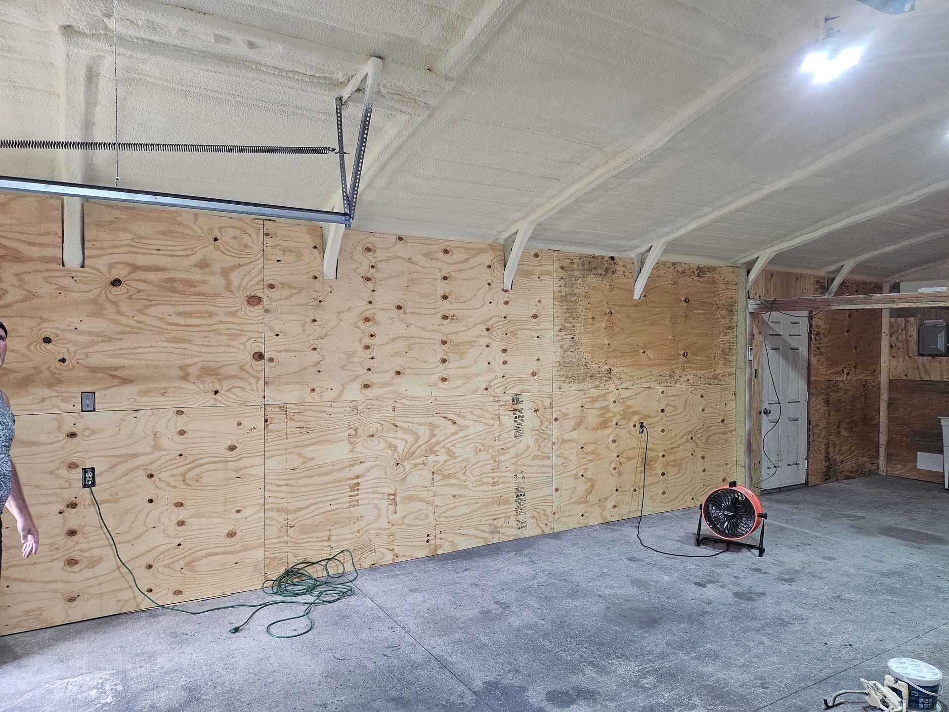 Garage interior with plywood walls, insulation on the ceiling, and a fan on the concrete floor.