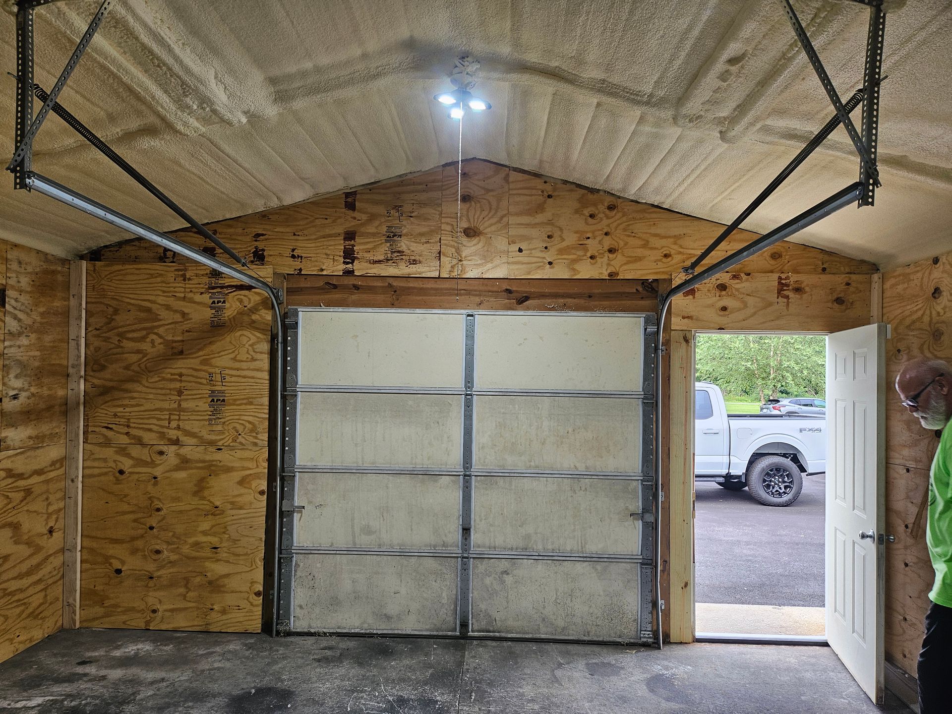 Inside a shed: garage door, open doorway with a truck, and a side door. Plywood walls and ceiling with foam insulation.