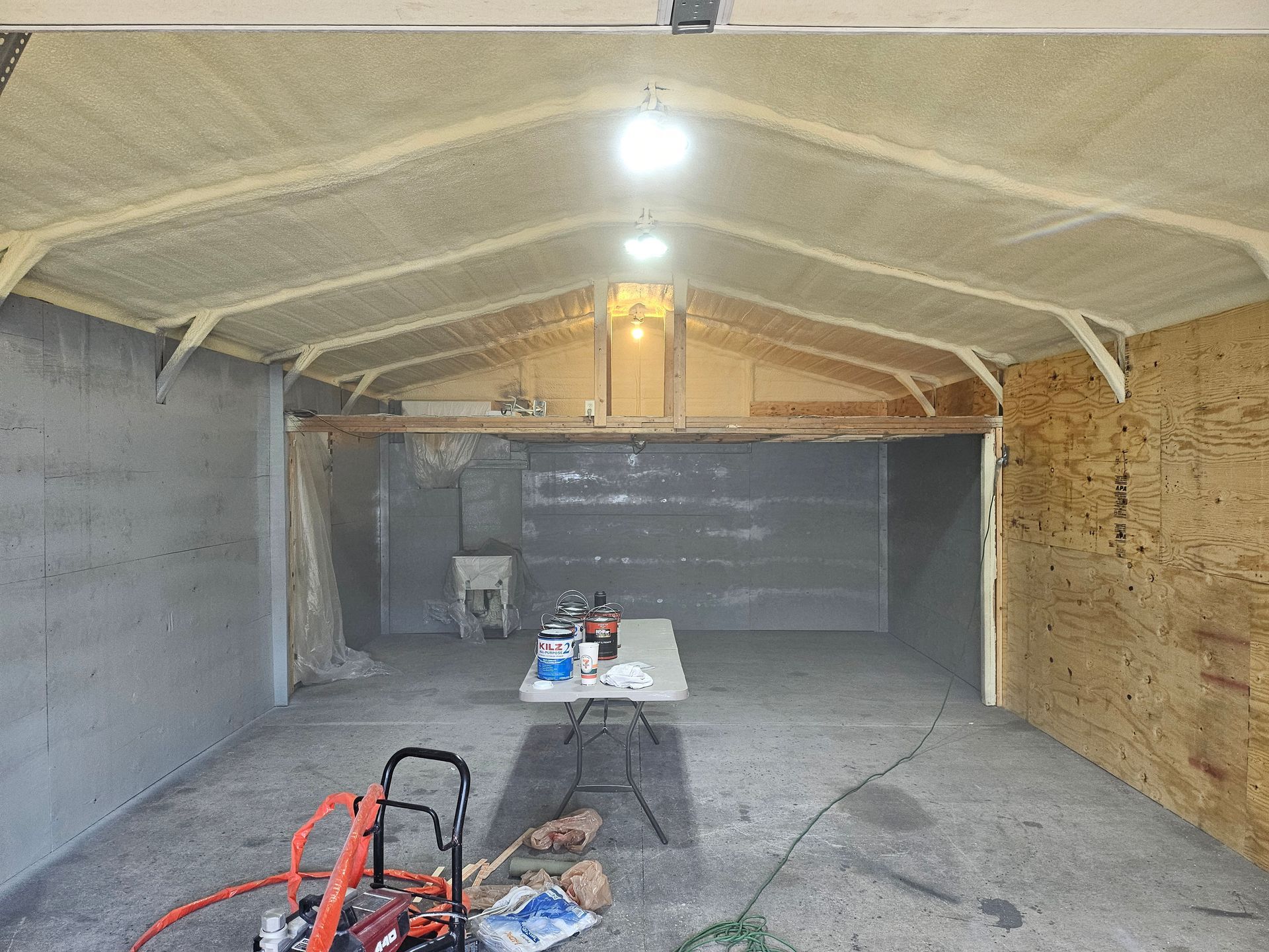 Interior of a garage with spray foam insulation, concrete walls, a table, and work equipment.