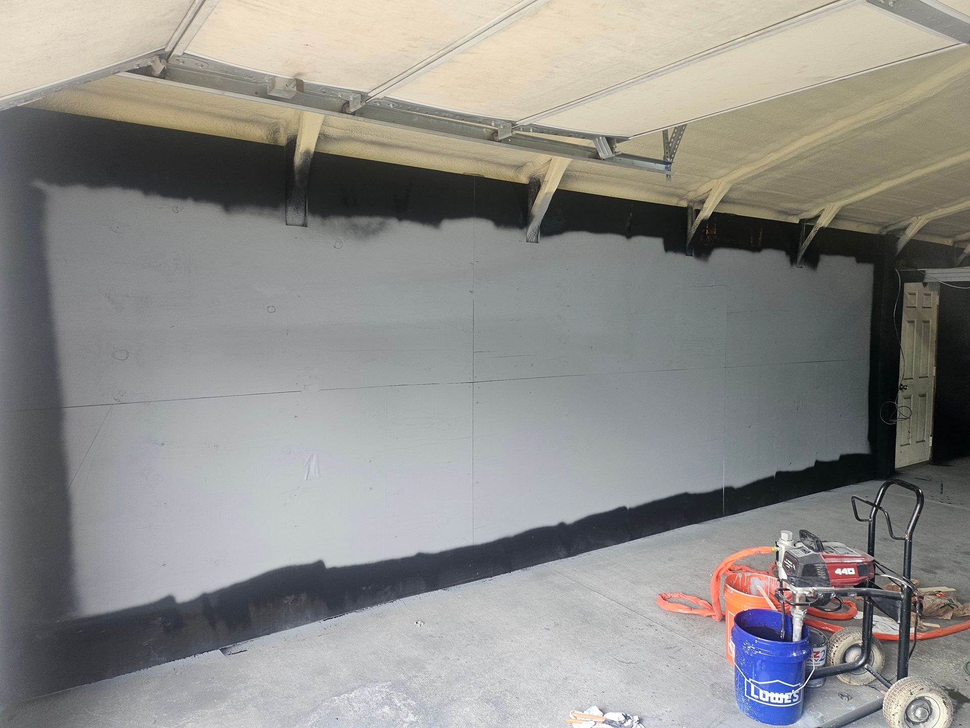 Interior wall being sprayed with gray insulation; equipment and blue bucket visible.