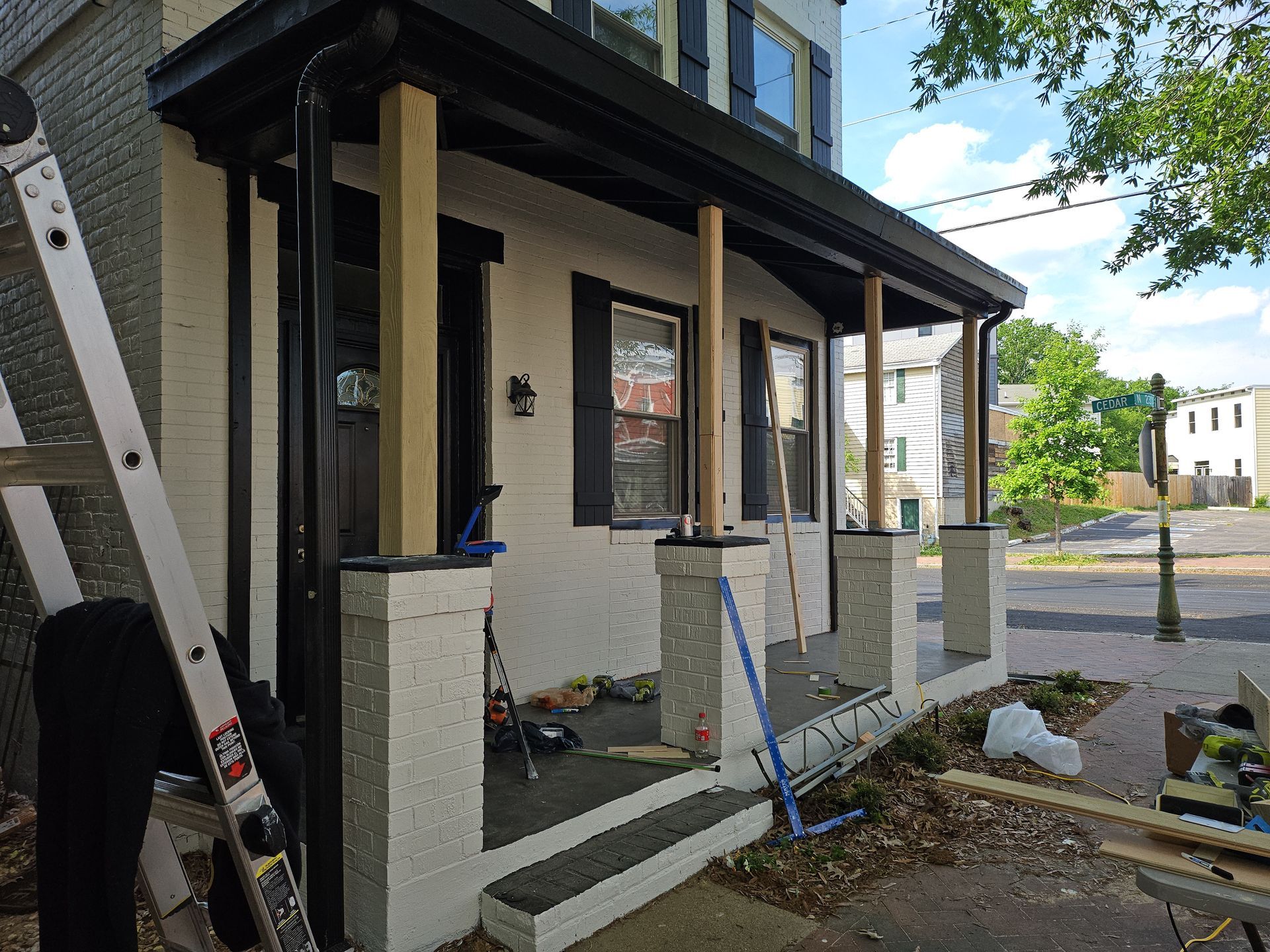 Porch under construction; brick house with white walls, black trim, wooden posts, and a ladder.