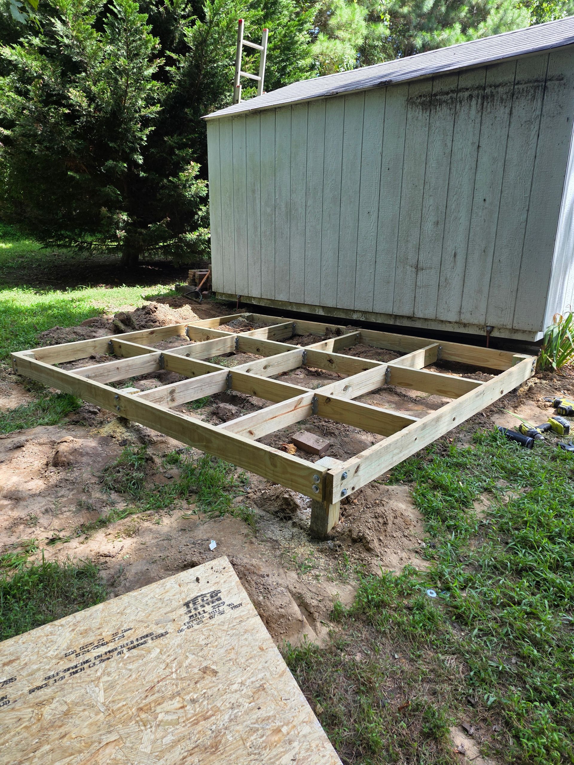 Wooden shed foundation being built, near a shed and a ladder, on a grassy area.