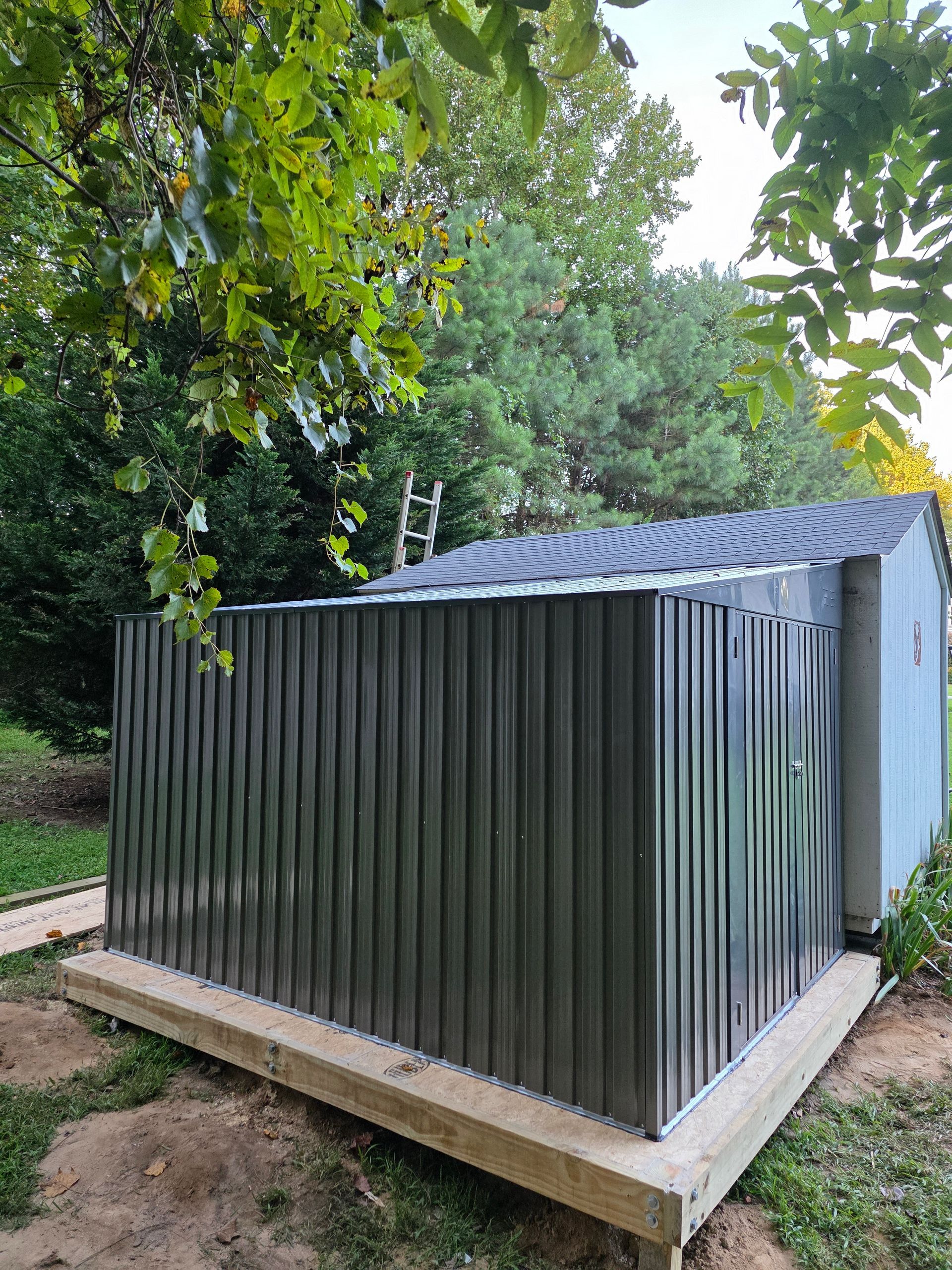 Metal storage shed with corrugated walls and a black roof, on a wooden base.