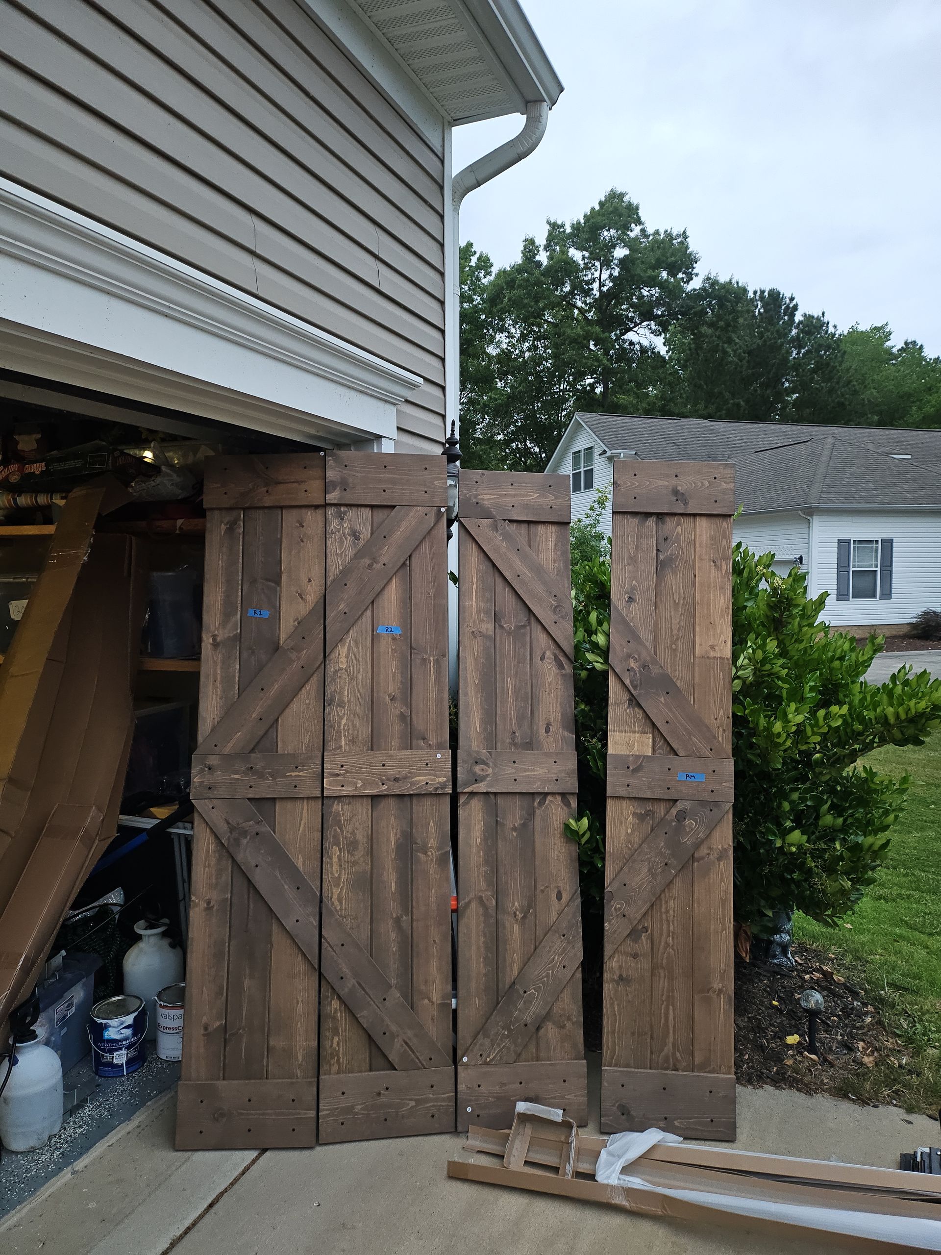 Three wooden barn doors leaning against a house exterior; brown with diagonal accents.