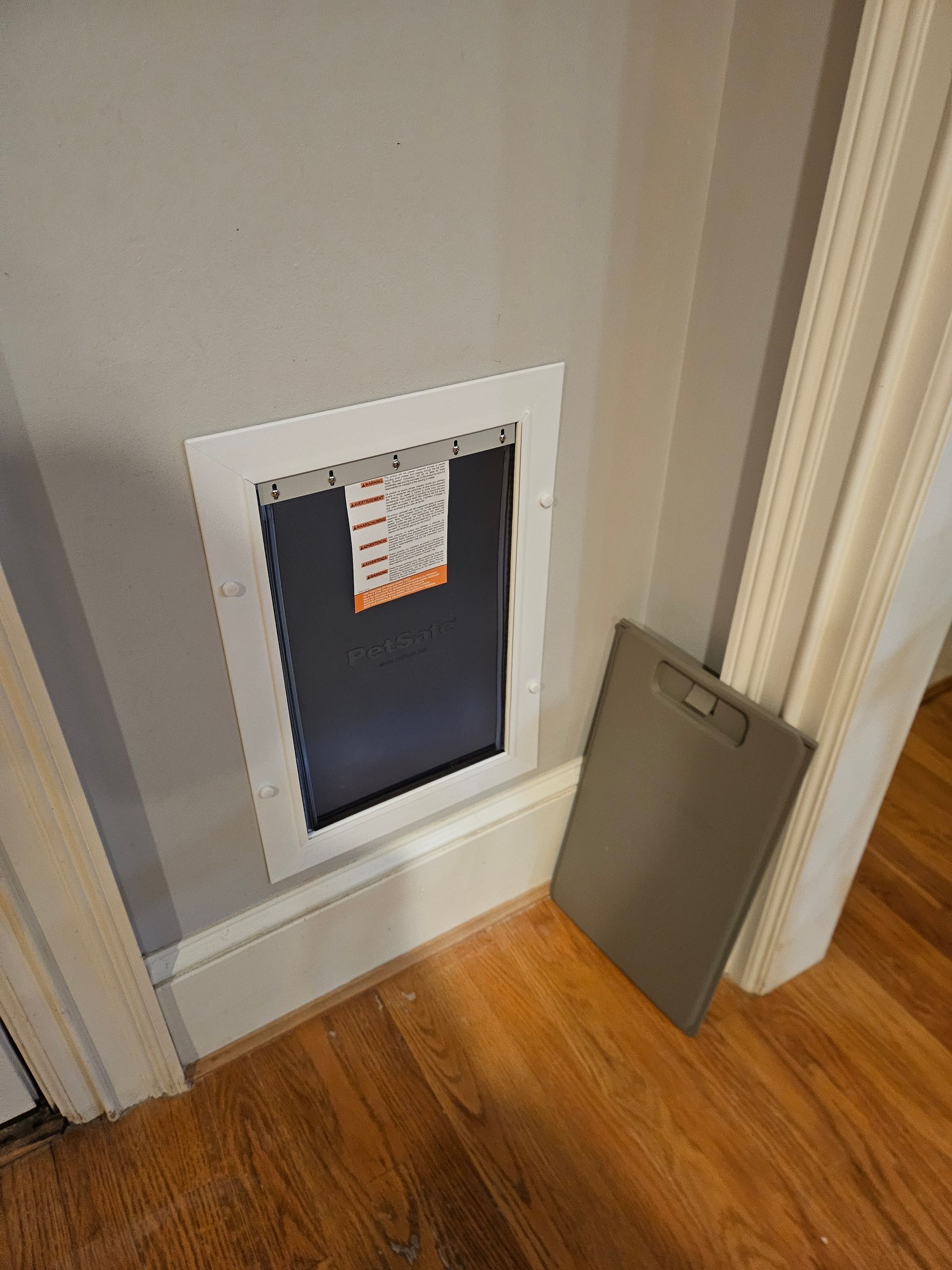 Dog door in a gray wall with a white frame, next to a closed, gray panel on a wood floor.