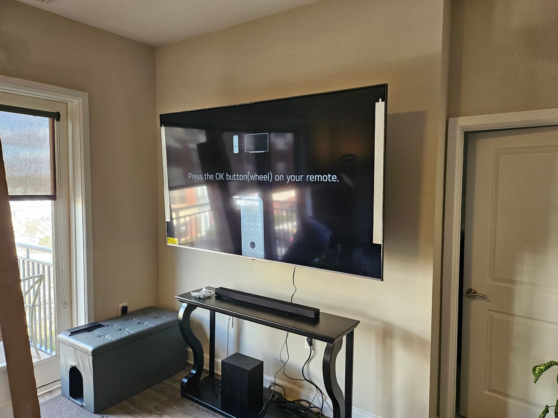 Large, wall-mounted TV over a console table. A soundbar and subwoofer are on the table. Beige walls, door, window.