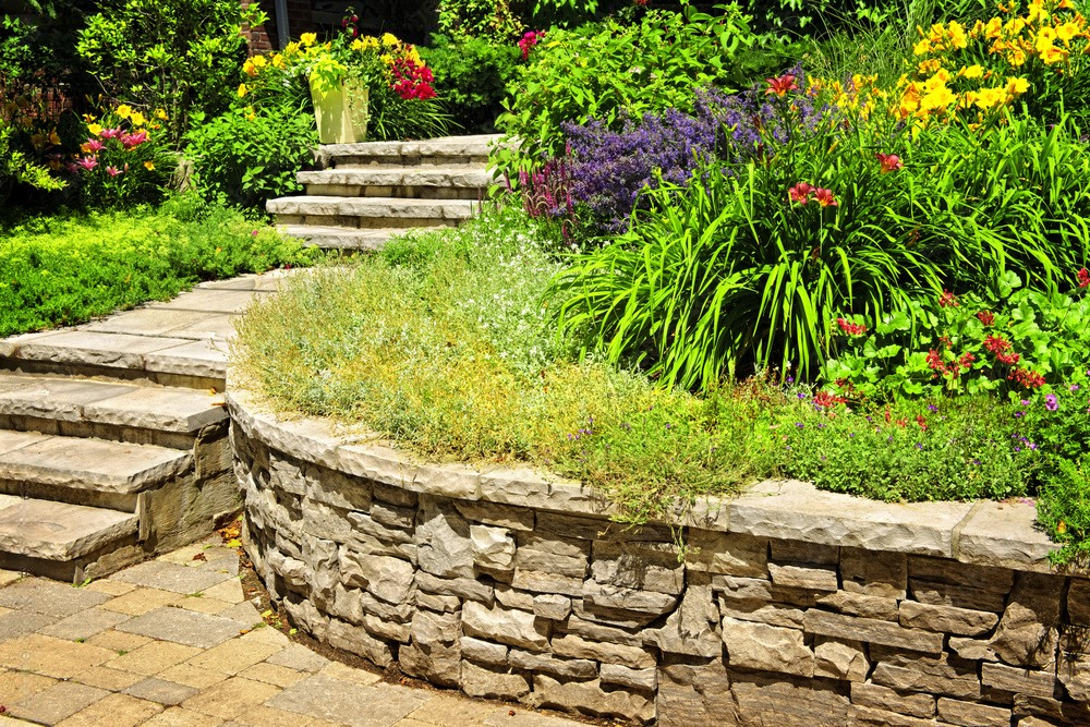 a garden with a stone wall and stairs surrounded by flowers