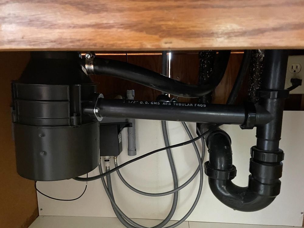 Under-sink plumbing with black drain pipes, garbage disposal, and visible cords against a white cabinet wall