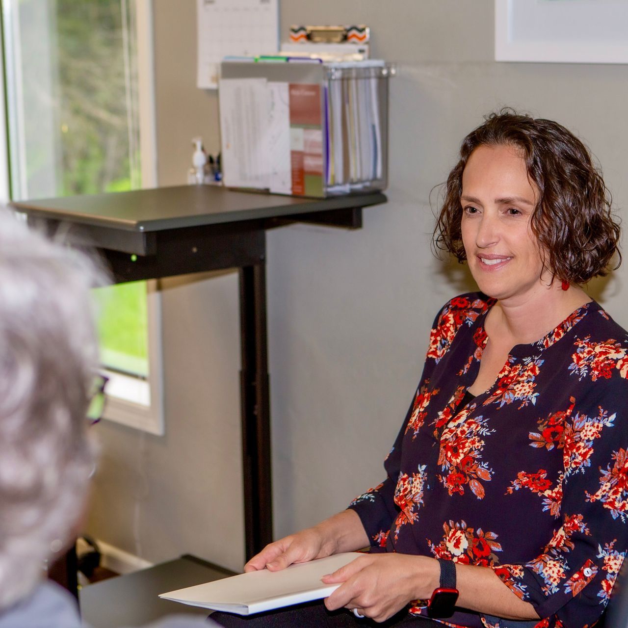 A woman in a floral shirt is sitting at a desk talking to another woman.