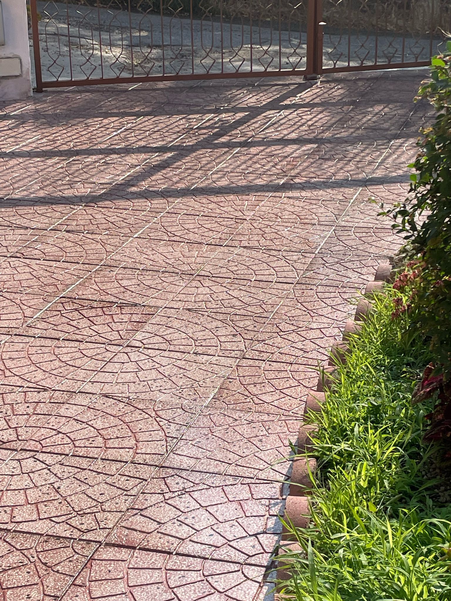 Vialetto d'accesso in mattoni decorativi con ombre provenienti da un cancello in ferro battuto. Piante verdi delimitano il lato destro.