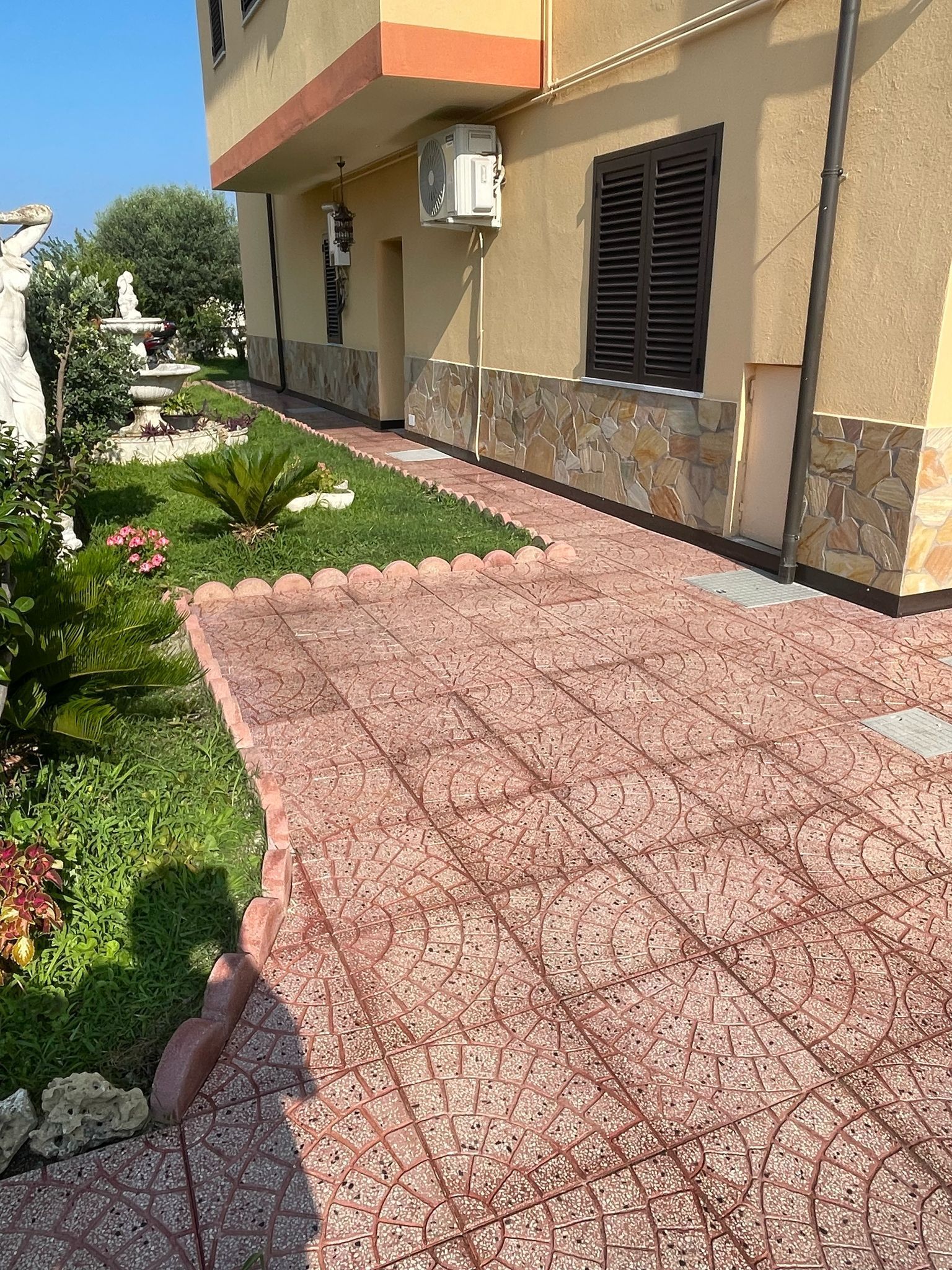 Un edificio con un vialetto in mattoni e un giardino. L'esterno è in stucco marrone chiaro e una finestra con persiane marroni.