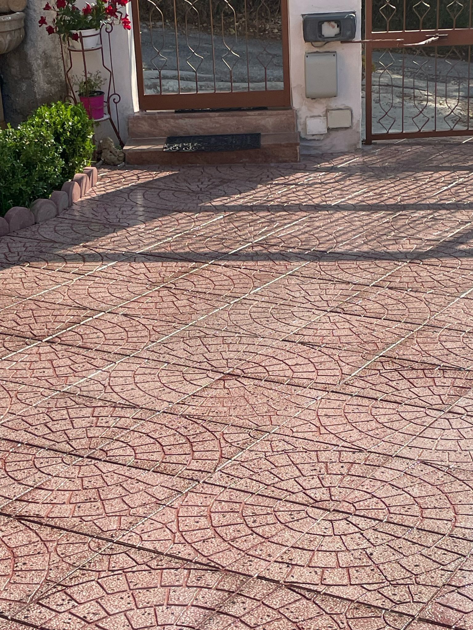 Area pavimentata in mattoni rossi che conduce a un cancello e a una casa. Un piccolo vaso di fiori e delle piante si trovano vicino all'ingresso.