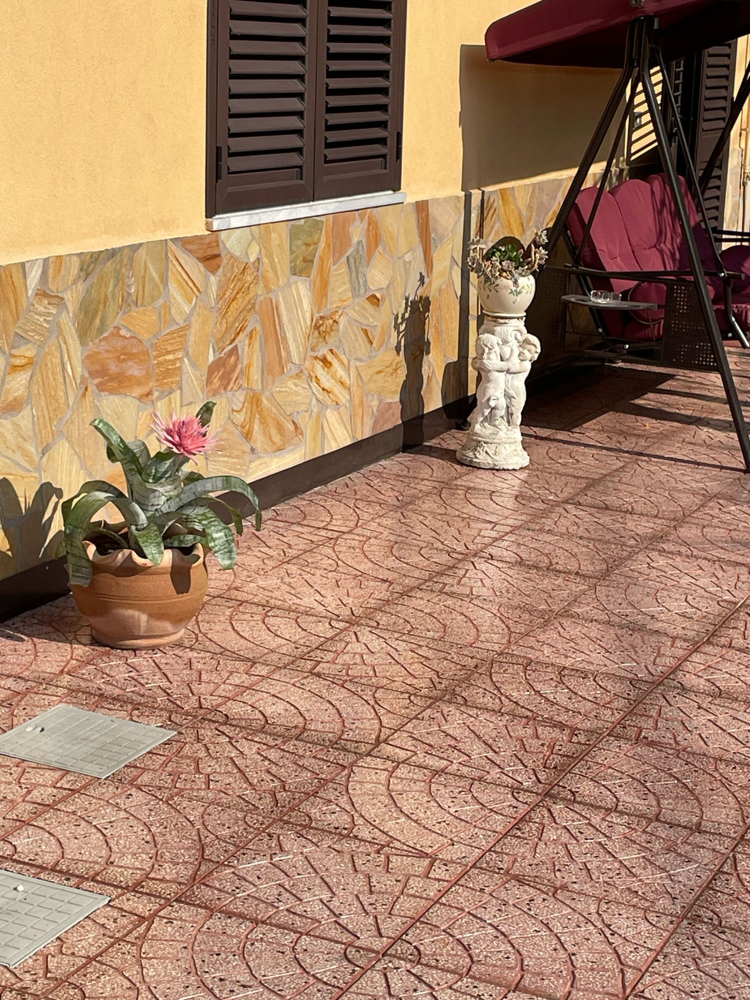Un patio in terracotta con piastrelle decorative, un vaso di fiori e un'altalena, accanto a un edificio con rivestimento in pietra.