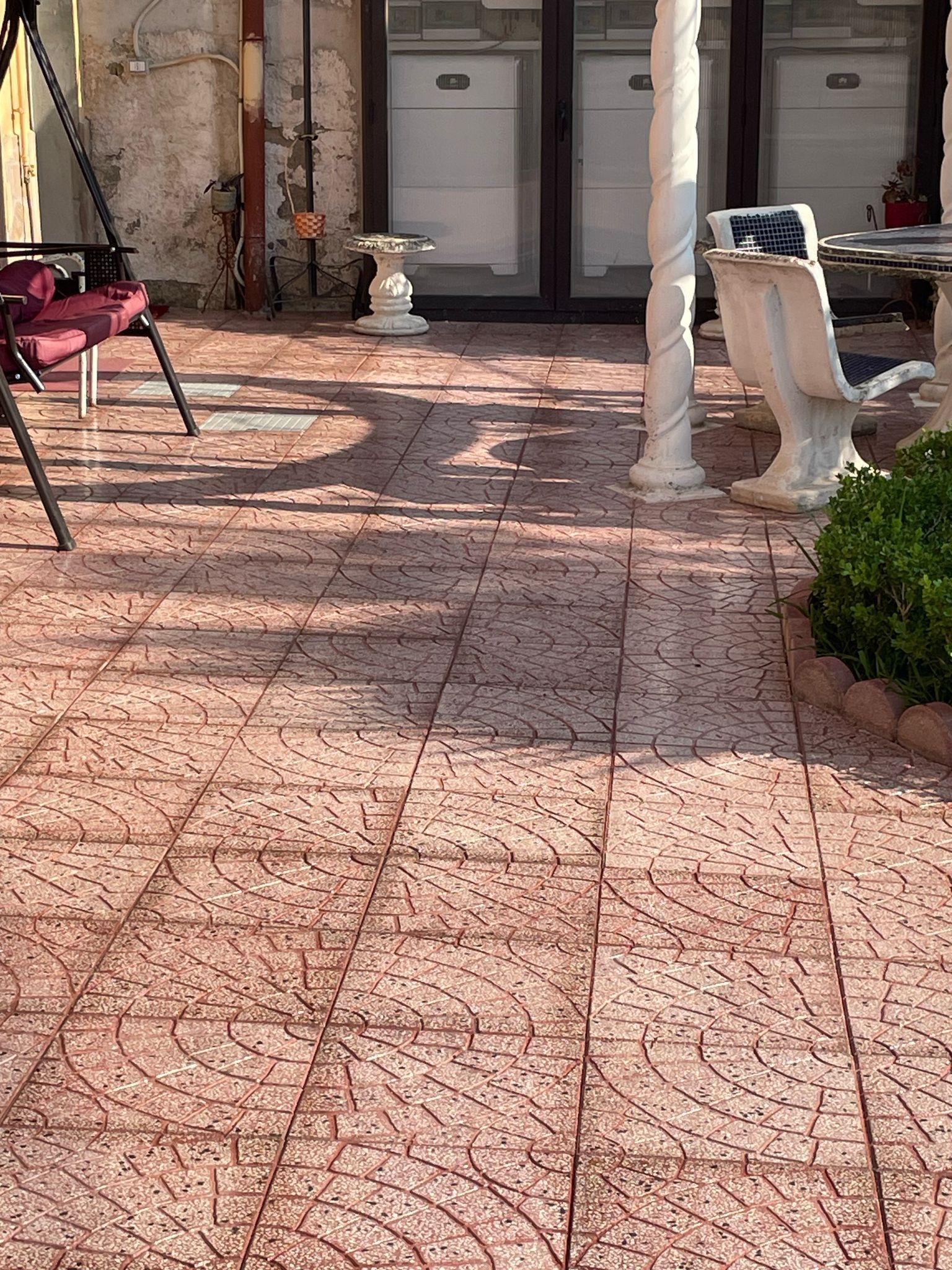 Patio terrazzato con piastrelle a motivi rosso-marroni, una colonna bianca e mobili da esterno sotto un cielo soleggiato.