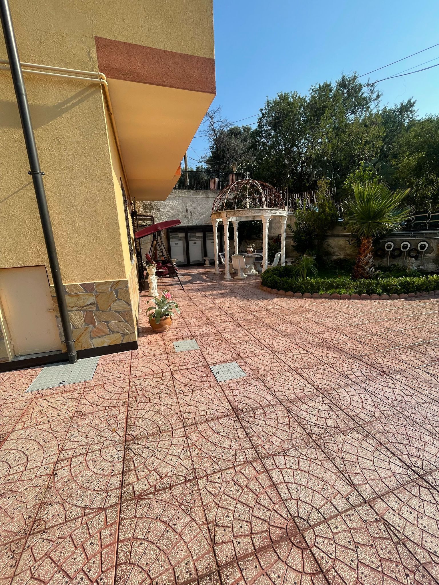 La facciata beige di un edificio con una piazza pavimentata in primo piano, che conduce a un gazebo bianco e a un giardino sotto un cielo azzurro e terso.