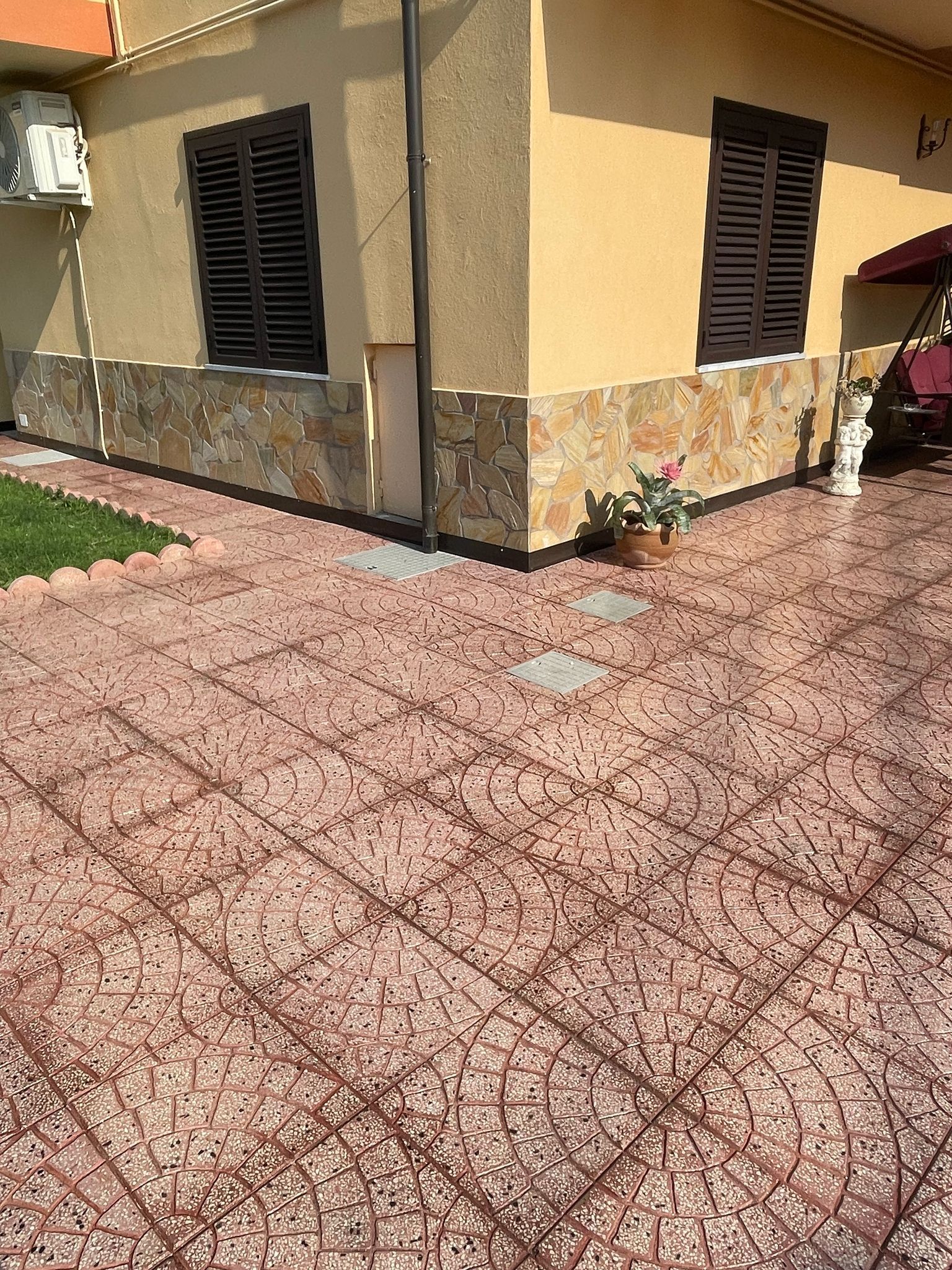 Vista esterna di una casa con persiane marroni, pareti color cuoio e un patio in mattoni decorati. Alla base della casa sono presenti pietre decorative.