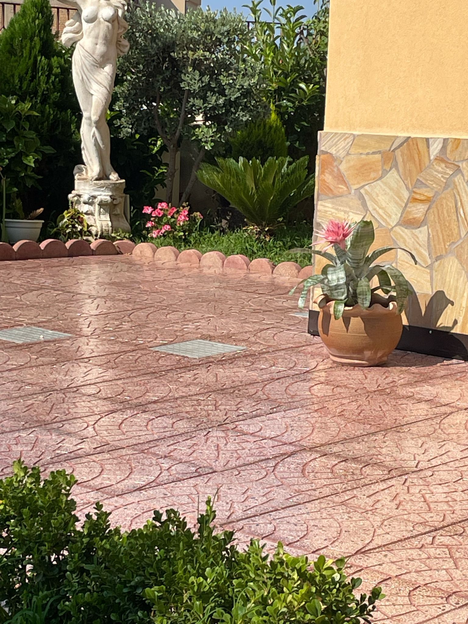 Un patio soleggiato con una superficie in cemento stampato color rosa. Una statua in pietra e piante in vaso sono inserite in un giardino.