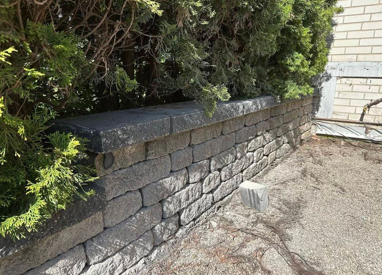 A retaining stone wall next to a brick building with a bush in the background.