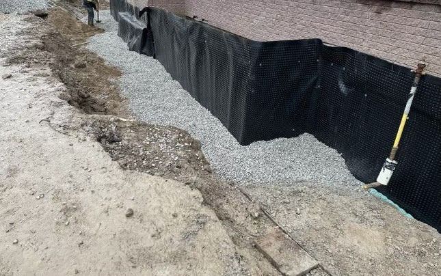 A damp-proofing membrane installed on the side of a home, showcasing a waterproofing service. The image also features a dug trench along the home's foundation, illustrating the process of protecting the home from water damage.