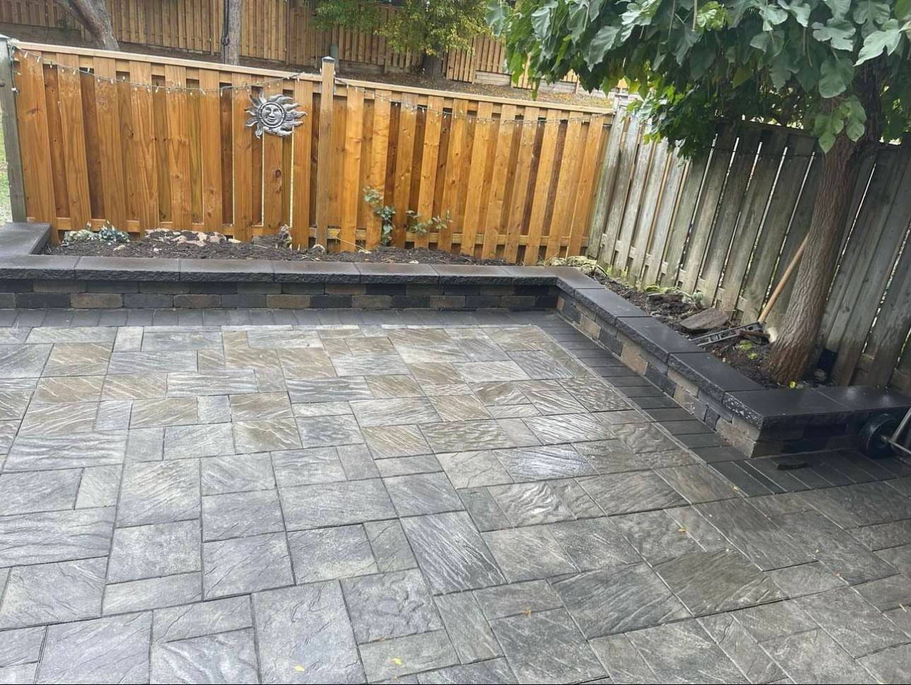 A jewel-stone patio with a wooden fence and a tree in the background.