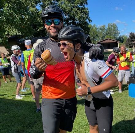 Two cyclists celebrating with a donut. One is holding the donut, the other is pretending to eat it. Outdoors, sunny.