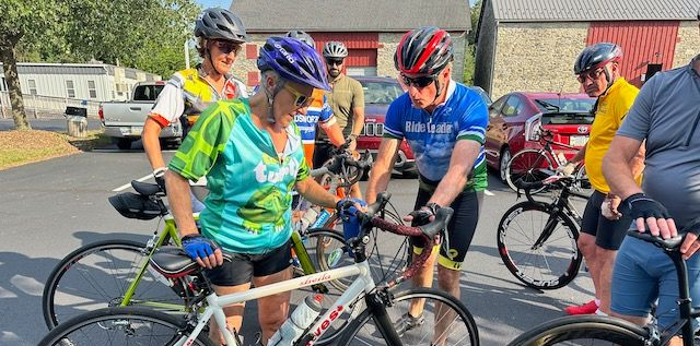 Cyclists gather around bikes, checking them. Outdoors with buildings and cars.