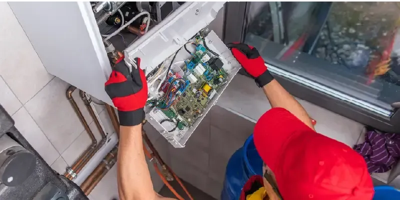 A person in a red hat and gloves works on a circuit board inside a heating unit near a window.