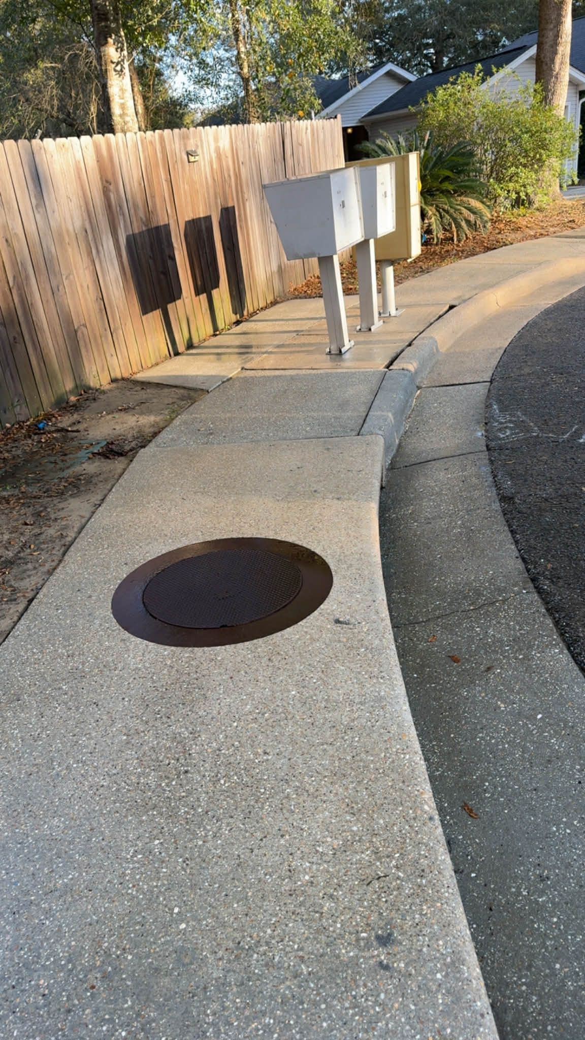 Concrete sidewalk with a manhole cover, mailbox, and wooden fence freshly pressure-washed.