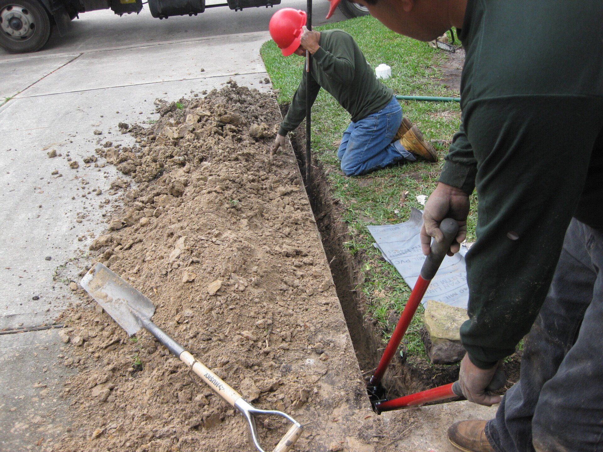 Two workers digging trench beside a road, one using shears, the other kneeling.