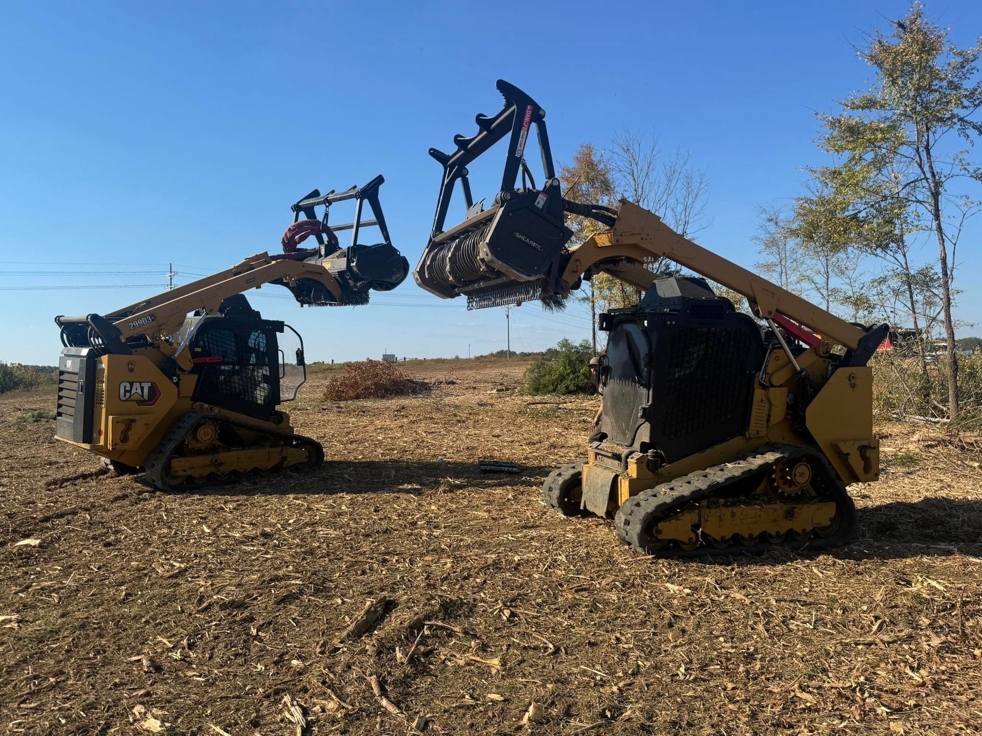 Two yellow skid steer loaders with forestry mulchers clearing a wooded area on a sunny day.