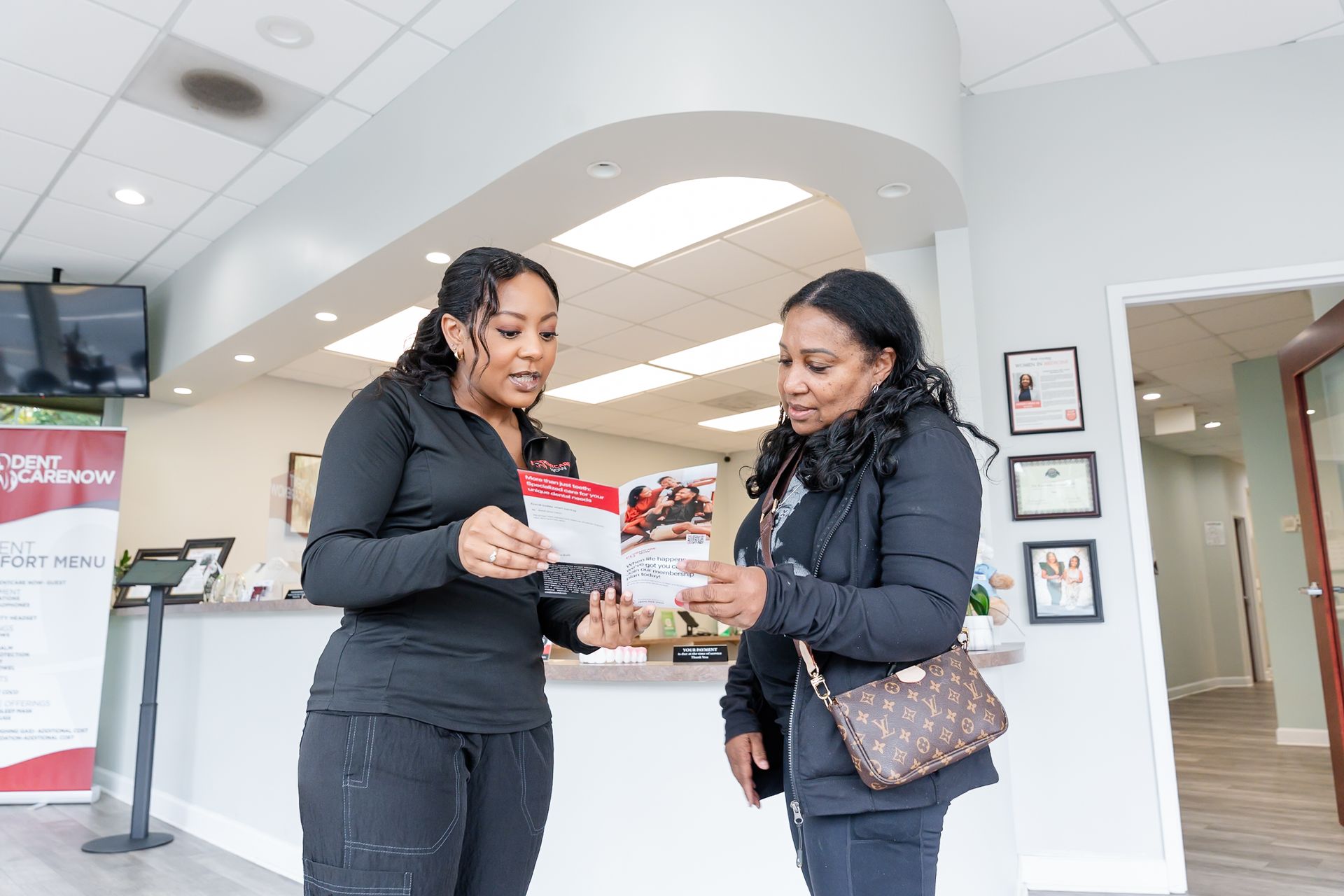 Two people in a bright office space discuss a promotional pamphlet held between them while standing at a reception desk.