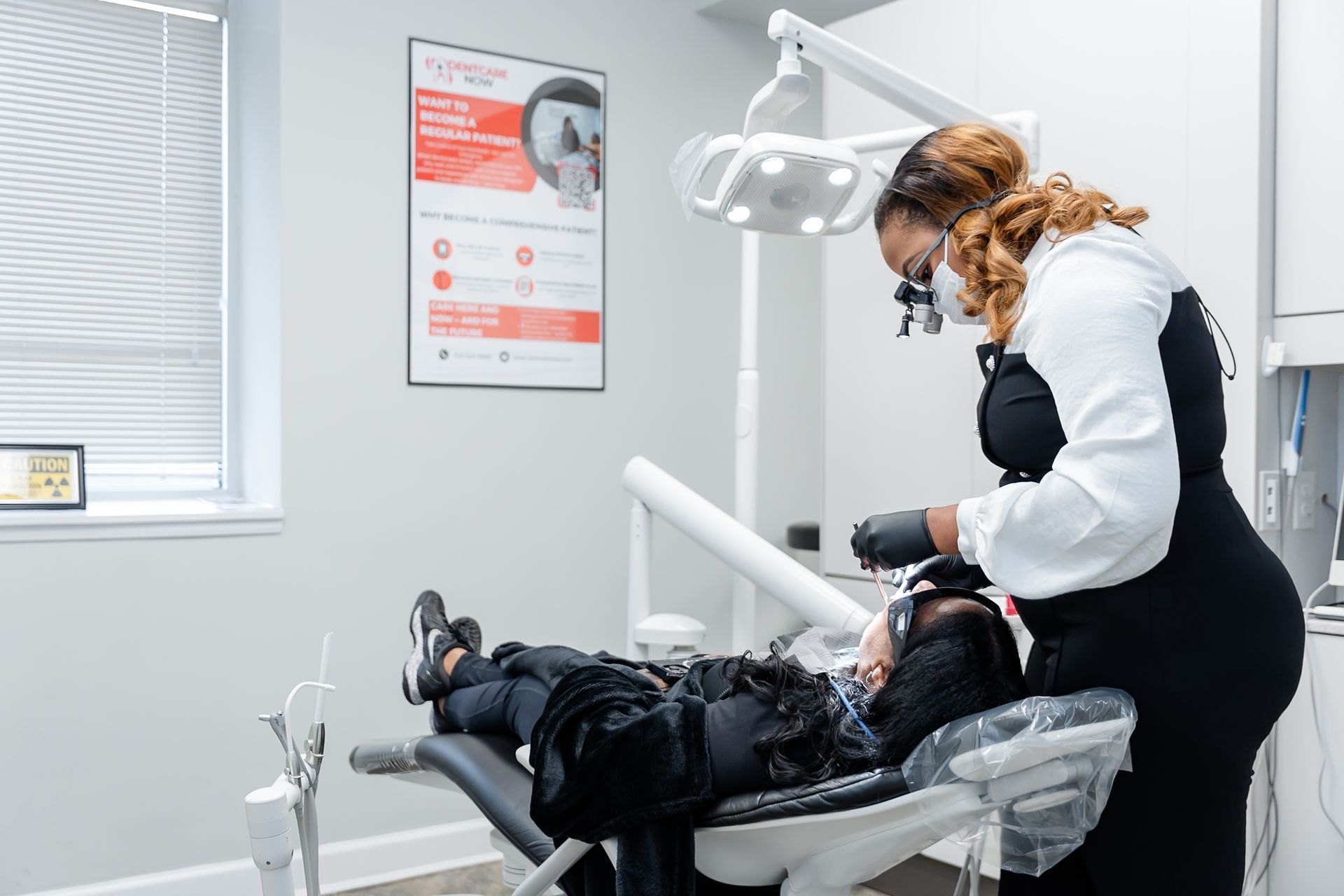 A dental professional wearing magnification loupes and a mask performs a procedure on a patient in a dental chair.