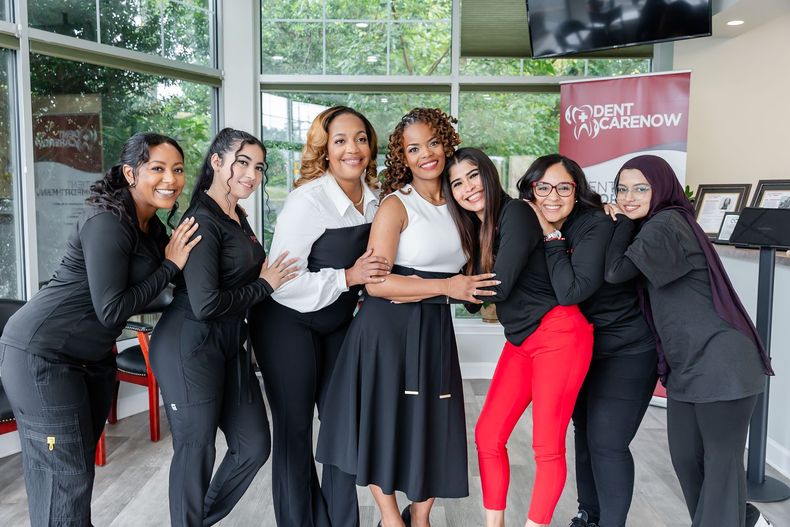 Seven smiling people pose in a bright, modern office with large windows, wearing professional attire and scrub sets.