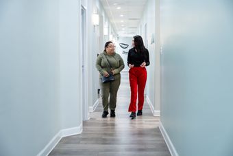 Two people in casual attire walk and talk down a bright, modern hallway with light blue walls and wood-patterned flooring.