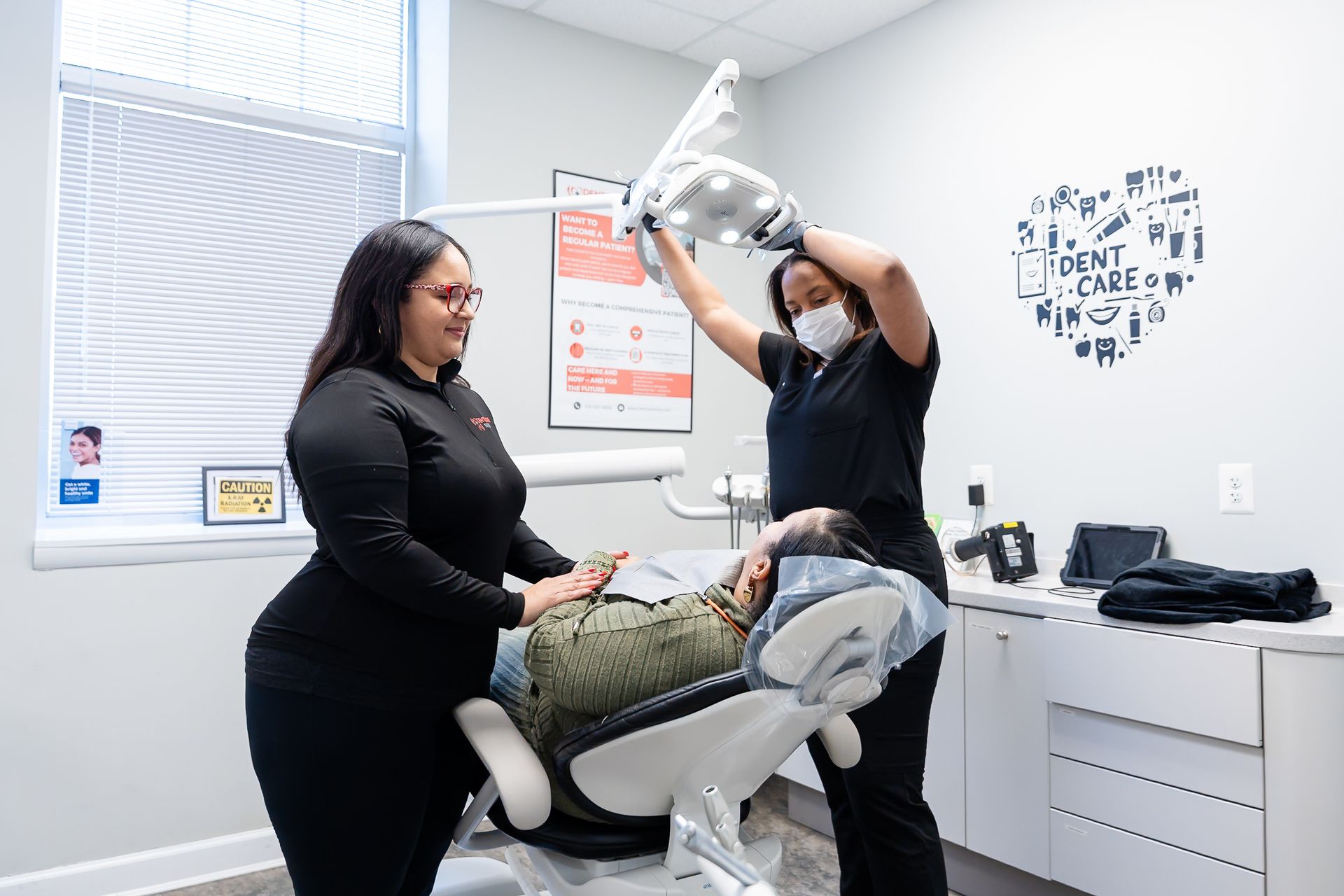 A dental professional adjusts a light over a patient in a dental exam chair while a colleague assists beside them.