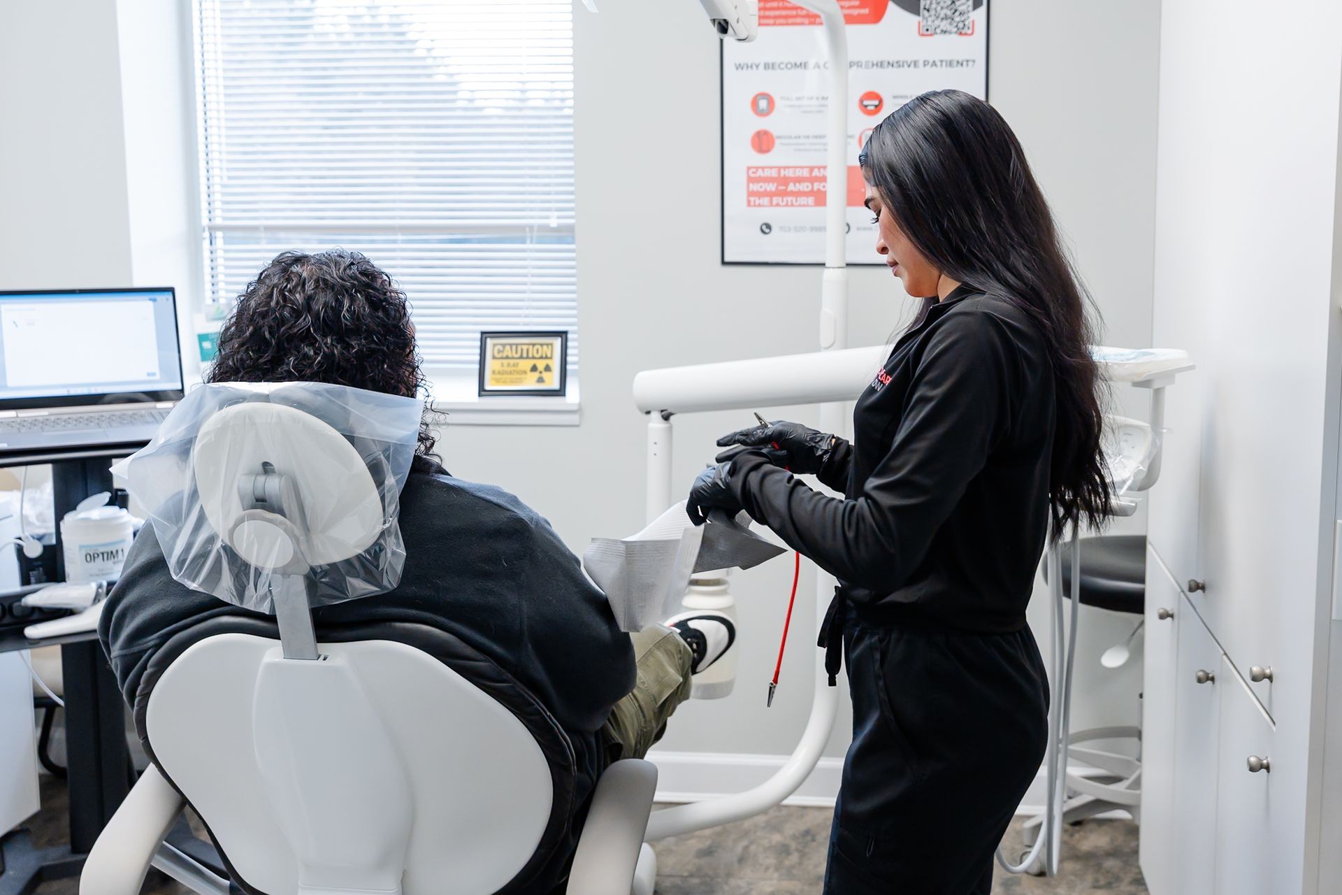 A professional in black attire confers with a patient seated in a dental chair within a clinic setting.