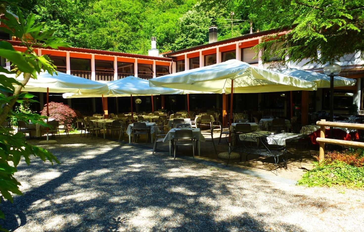 Patio del ristorante con tavoli e sedie, un edificio con finiture in legno e alberi sullo sfondo. Giornata di sole.