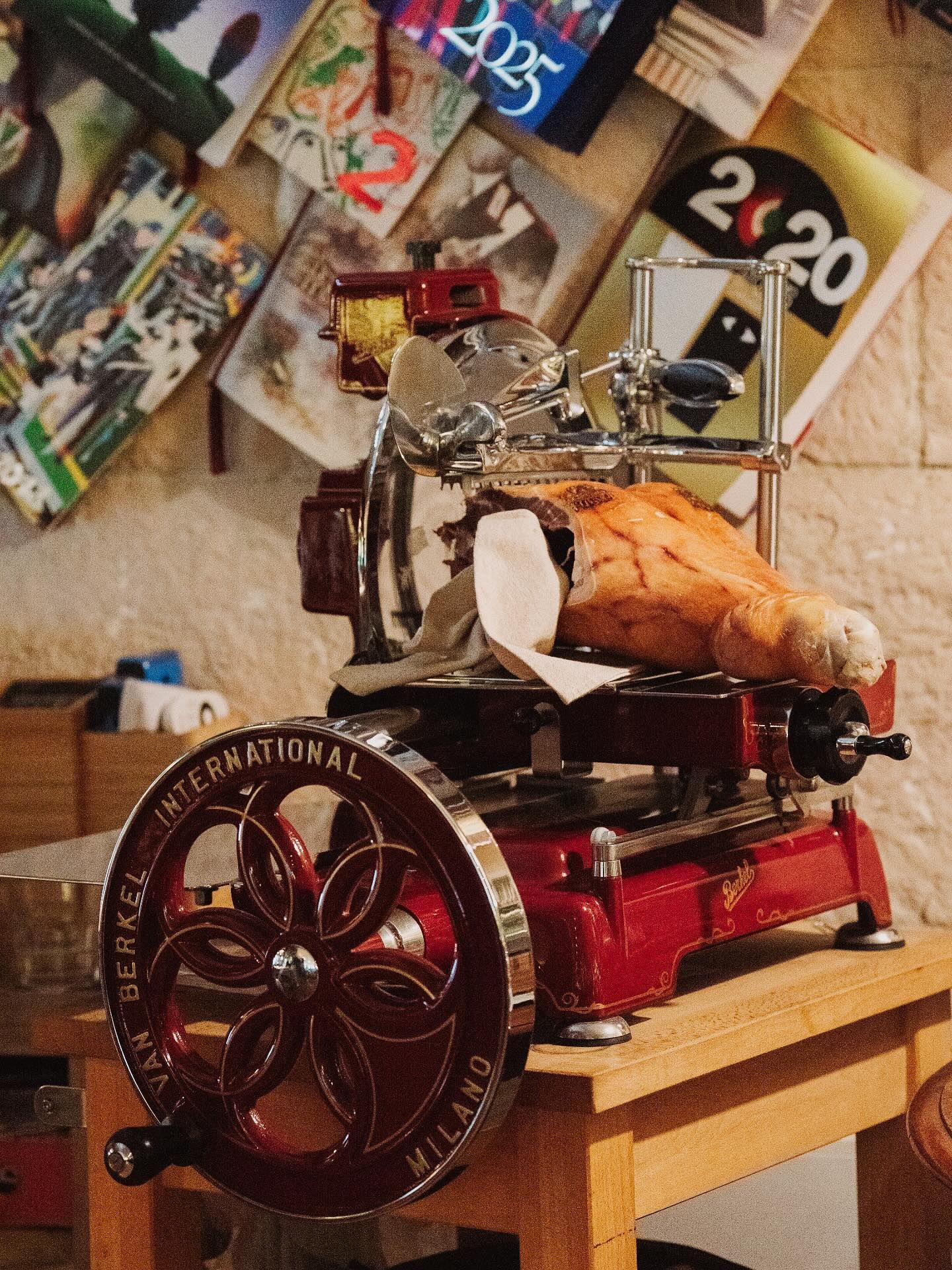 Affettatrice per carne rossa, che affetta parzialmente un grosso prosciutto, con la scritta "Sirman International Milano" sulla ruota; contro una parete con dei poster.