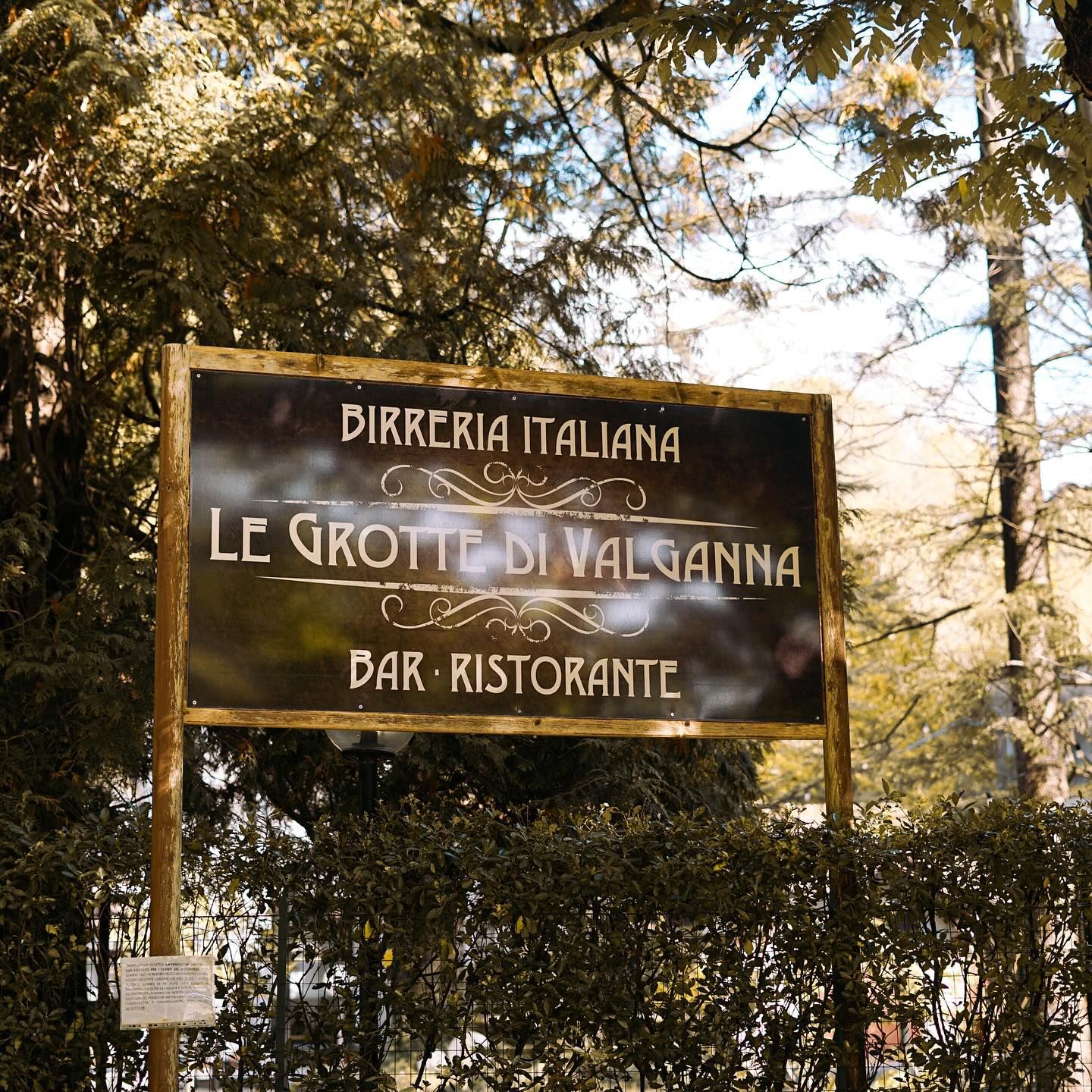 Insegna per il ristorante italiano "Le Grotte di Valganna" in una zona boscosa. Insegna nera con testo bianco.