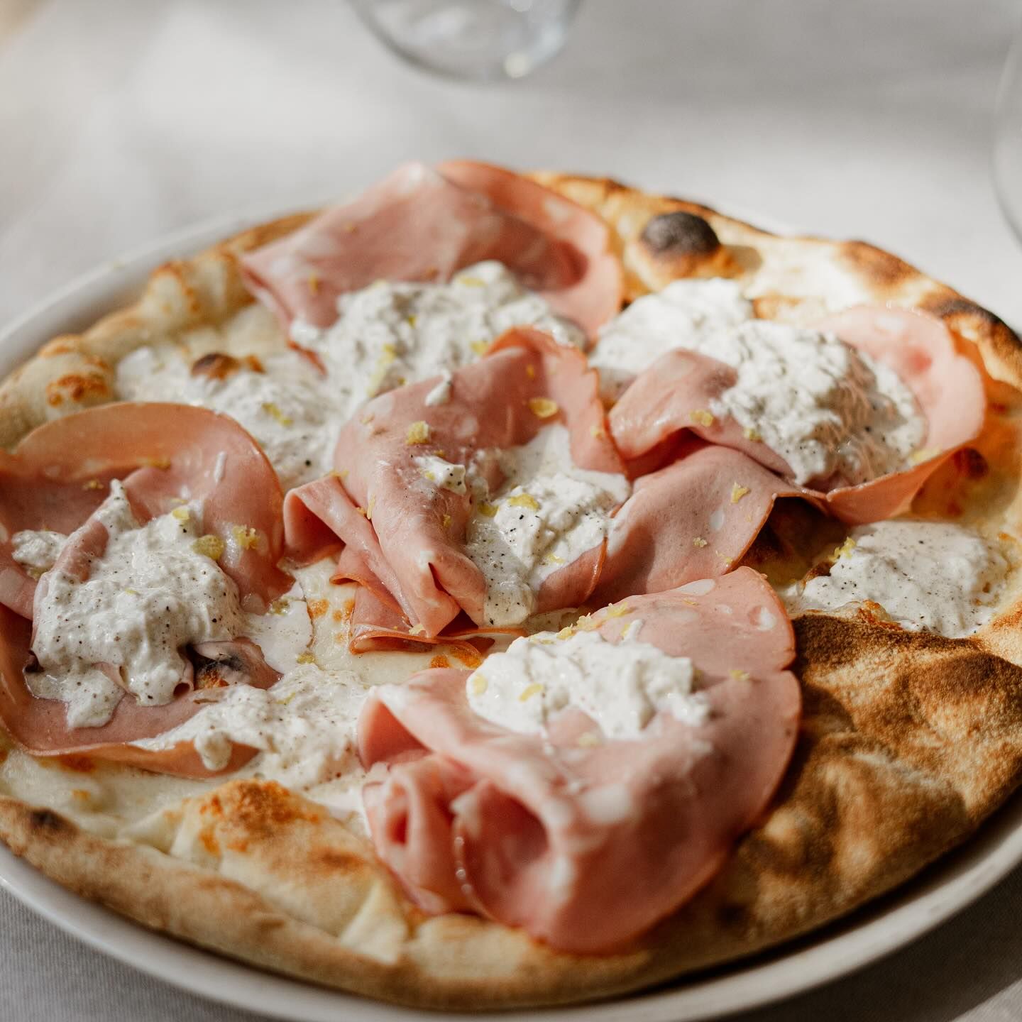Pizza condita con mortadella, formaggio cremoso e una leggera spolverata di scorza di limone su un piatto bianco.