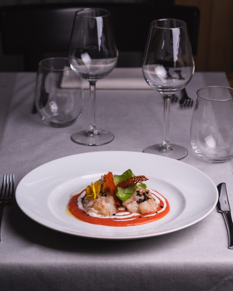 Piatto di cibo su un tavolo apparecchiato con bicchieri da vino e posate. Il cibo comprende frutti di mare con salsa rossa e verdure.