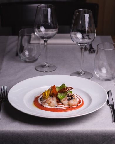 Piatto di cibo su un tavolo apparecchiato con bicchieri da vino e posate. Il cibo comprende frutti di mare con salsa rossa e verdure.