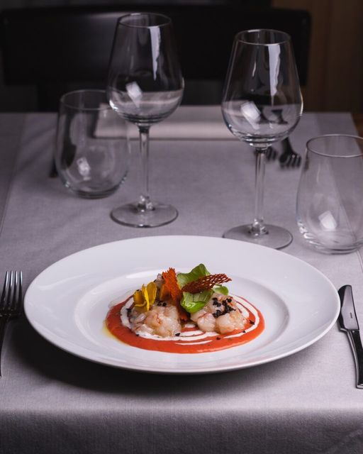 Piatto di cibo su un tavolo apparecchiato con bicchieri da vino e posate. Il cibo comprende frutti di mare con salsa rossa e verdure.