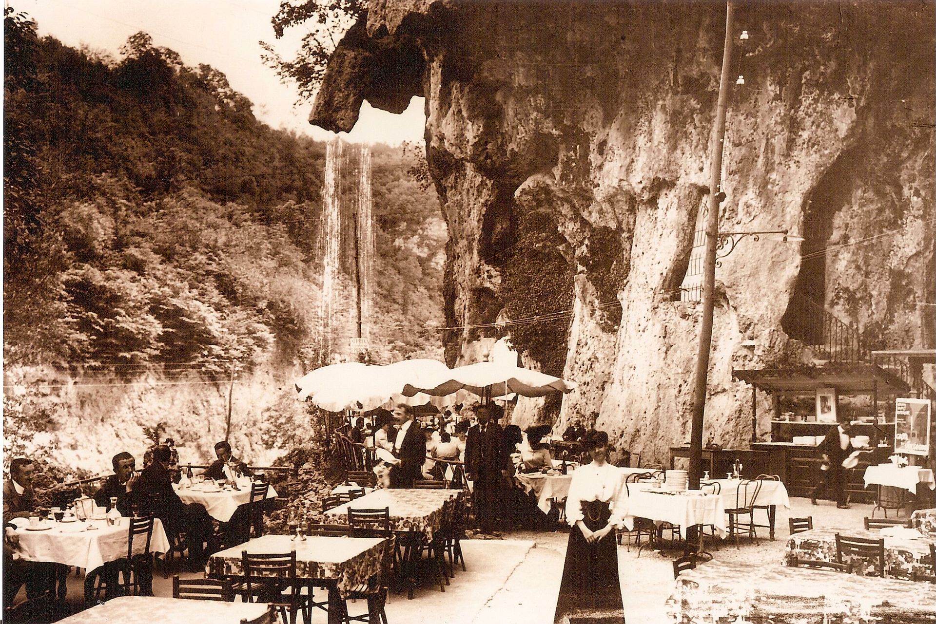Patio del ristorante con tavoli e sedie, un edificio con finiture in legno e alberi sullo sfondo. Giornata di sole.