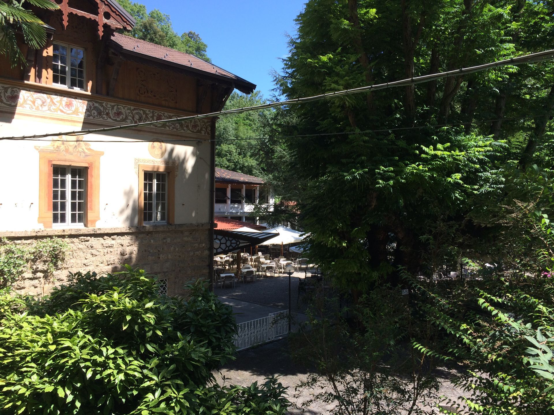 Esterno dell'edificio con zona pranzo all'aperto. Gli alberi incorniciano la scena sotto un cielo luminoso.