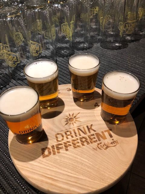 Quattro campioni di birra su un sottobicchiere di legno con la scritta "Drink Different" di Carlsberg. Sullo sfondo, bicchieri vuoti.