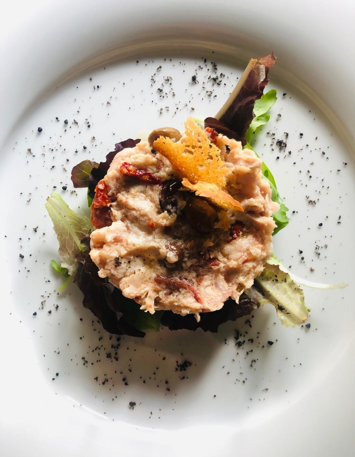 Antipasto di insalata di tonno su un letto di lattuga, guarnito con pomodori secchi, olive e un cracker su un piatto bianco.